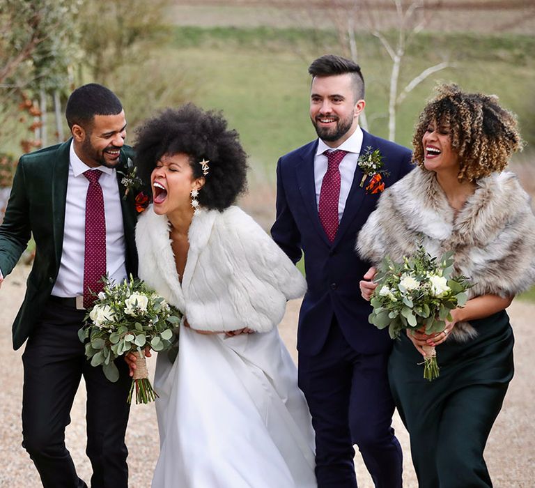 Bride & groom laugh with their wedding party as bride wears faux fur shawl outdoors and carries floral bouquet