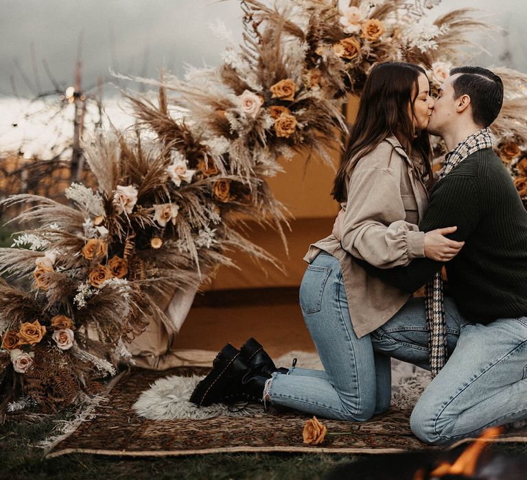 groom-to-be kissing his fiancé at their boho wedding proposal with bell tent and dried flower decor 