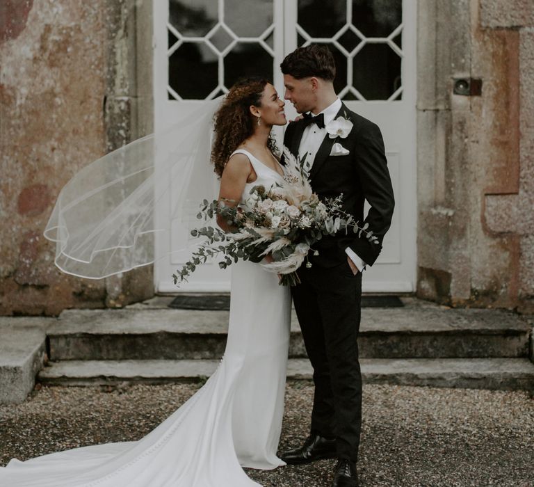 Bride & groom stand outdoors as brides veil blows in the wind