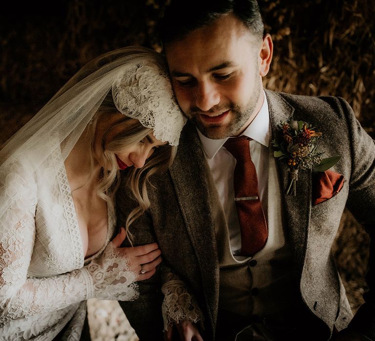 Bride in a lace wedding dress and Juliet cap veil leaning on her husbands shoulder in a brown wool suit