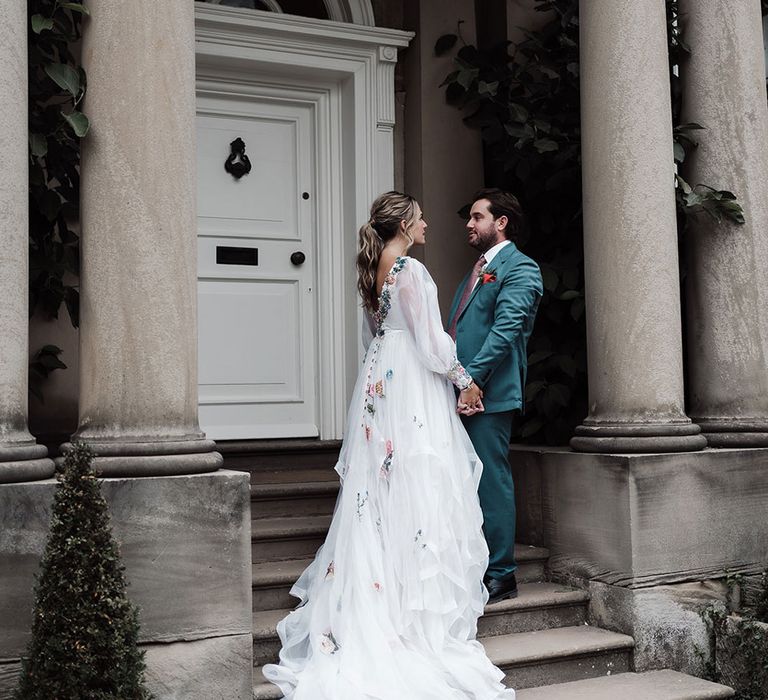 Bride in low back long sleeve embroidered wedding dress with train stands on stone steps holding hands with groom in teal suit