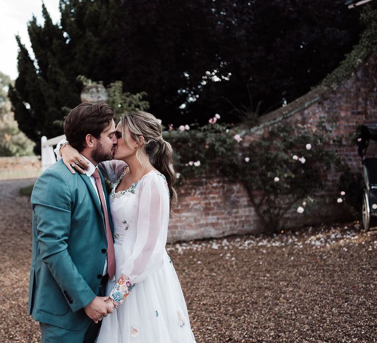 Groom in blue suit and pink tie kisses bride in long sleeve Victoria Sanders wedding dress with embroidered cuffs in the grounds of Iscoyd Park