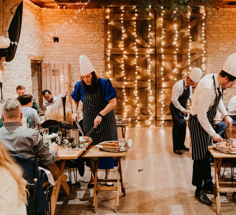 Guests in striped aprons and chefs hats cut up meats at tables for barn wedding reception with fairy light canopy