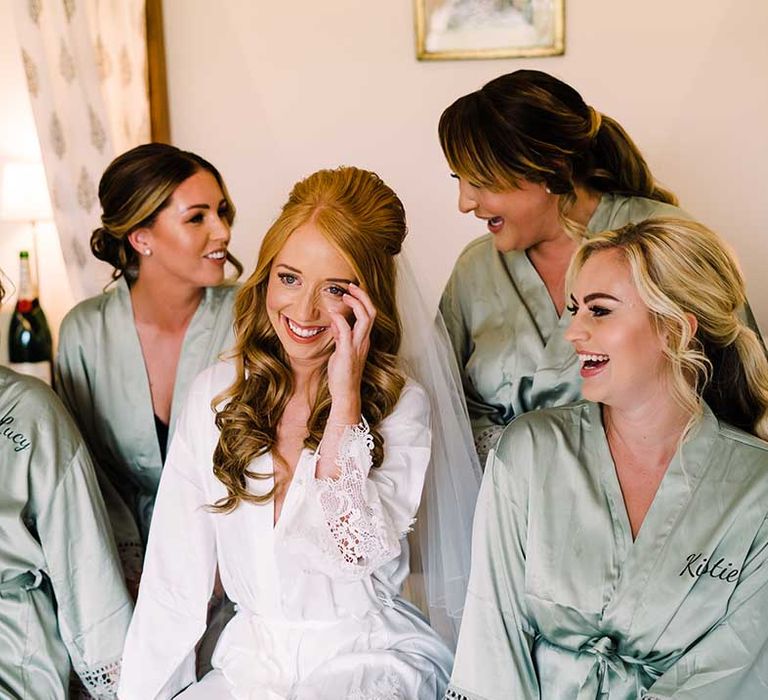 Bride laughs on her wedding day as bridesmaids wearing matching sage dressing gowns