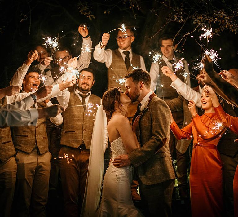 Bride and groom kiss under sparklers held by guests in burnt orange bridesmaid dresses