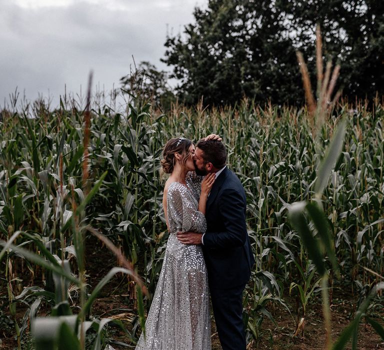 Bride in a silver sequin wedding dress and groom in a navy suit kissing amongst the green crops 