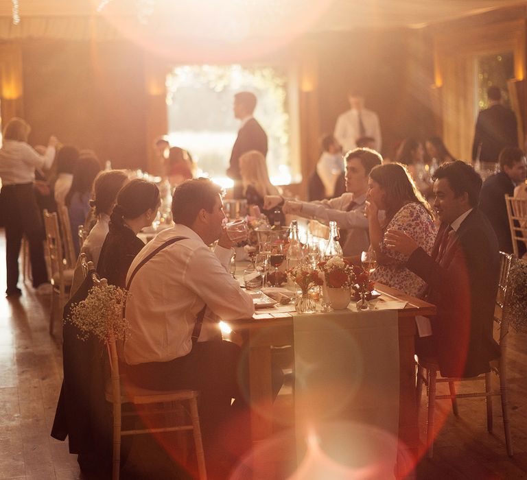 Wedding guests sit on bamboo chairs at long tables at Elmore Court wedding breakfast during golden hour