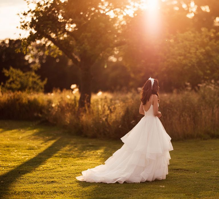 Bride in layered tulle skirt wedding dress with train and low back walks through grounds at Elmore Court during golden hour