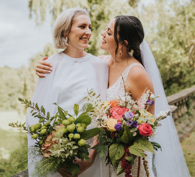 Bride in lace wedding dress hugging bride in jumpsuit with cape holding wedding bouquets 