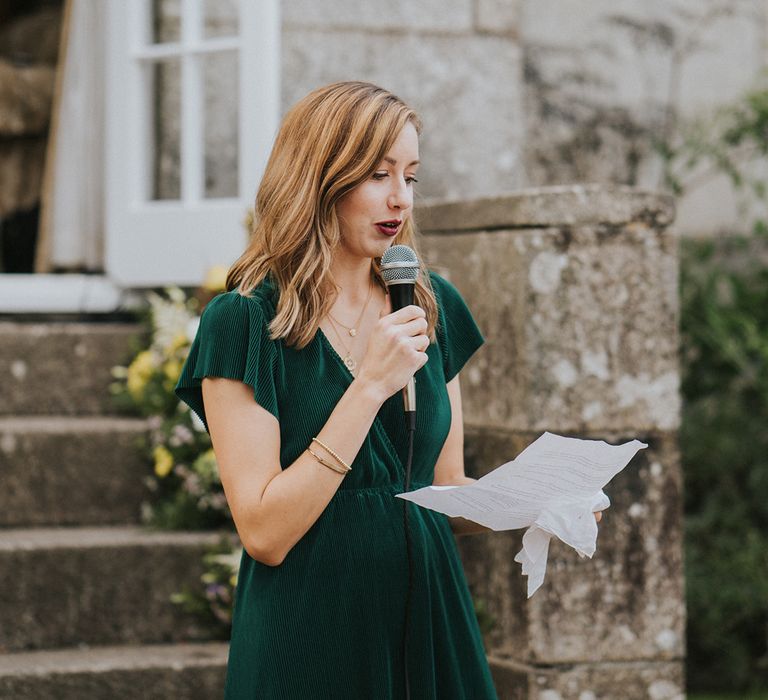 Bridesmaid in deep green dress reading out speech to the wedding party at country house venue 