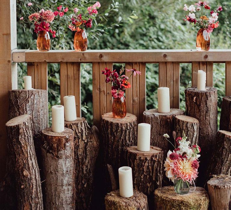 Wedding decor of different sized tree trunks with white pillar candles and colourful flowers in orange glass vases