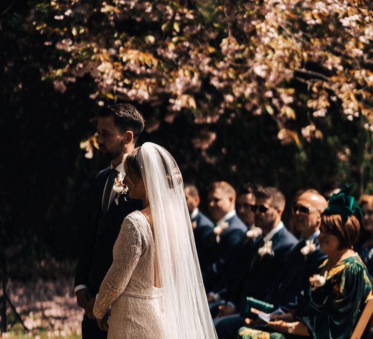 Beaded wedding dress with low back worn by bride in pearl veil with groom in dark blue suit at the altar 