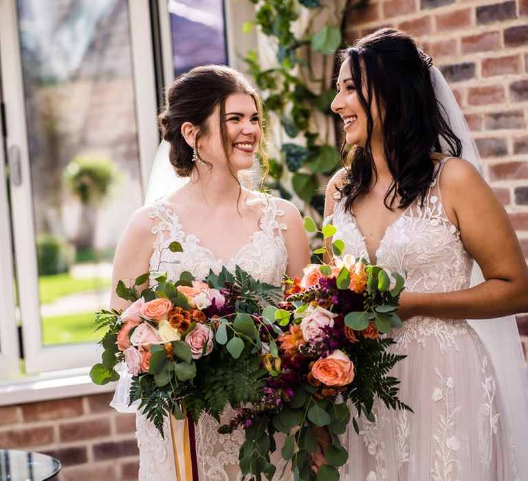 Brides in floral lace dresses with wedding bouquets with purple, orange and pink flowers include roses and fern leaves 