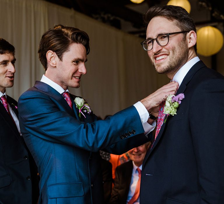 Groom in blue suit helps a groomsman with his matching pink and blue patterned tie