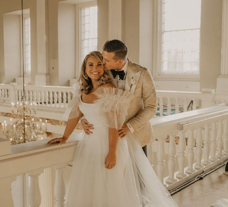 Groom in gold custom tuxedo kisses bride on her cheek wearing ruffle wedding dress and gold jewellery 