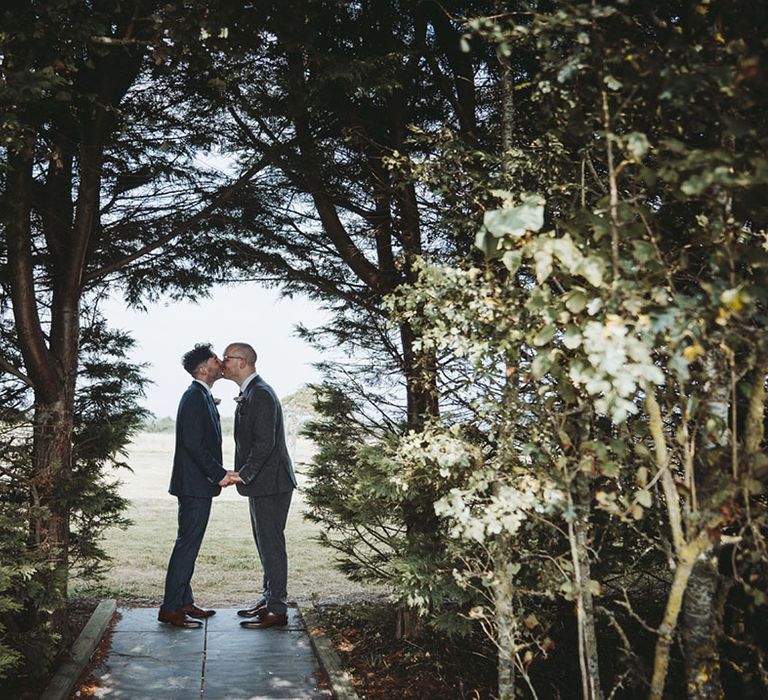 Grooms in grey and blue suits kiss each other as they stand under trees after their wedding