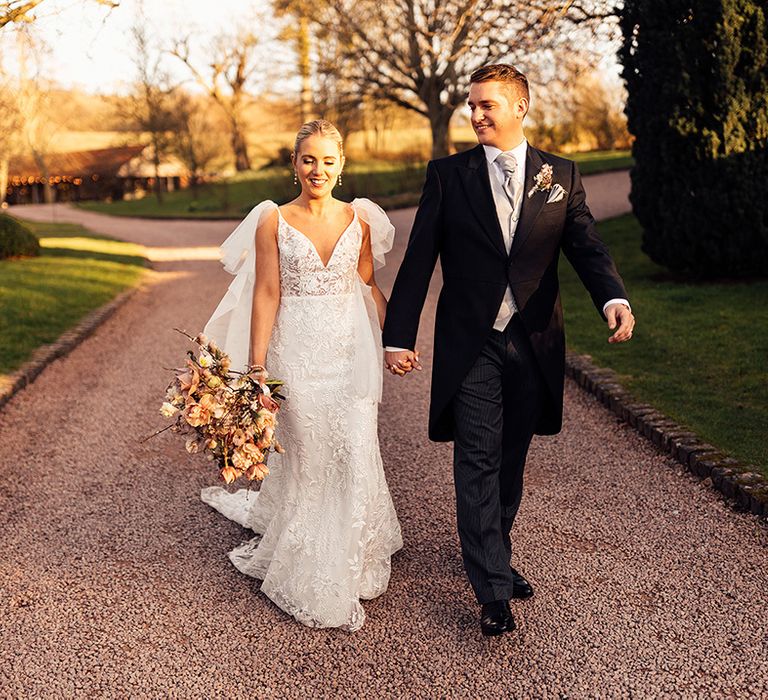 Bride holding neutral wedding bouquet walks around with groom in morning suit