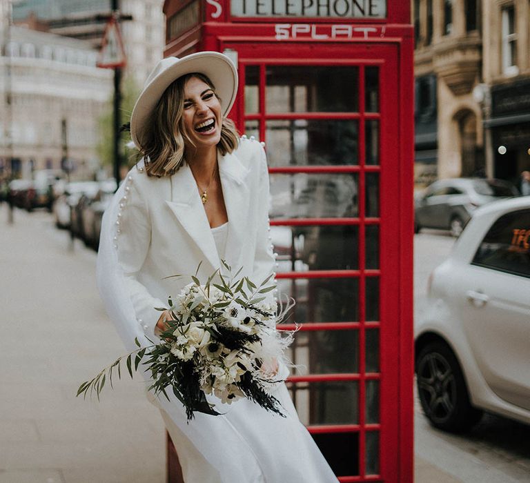 Bride laughing wearing a fedora and wedding suit with pearls with white flower bouquet