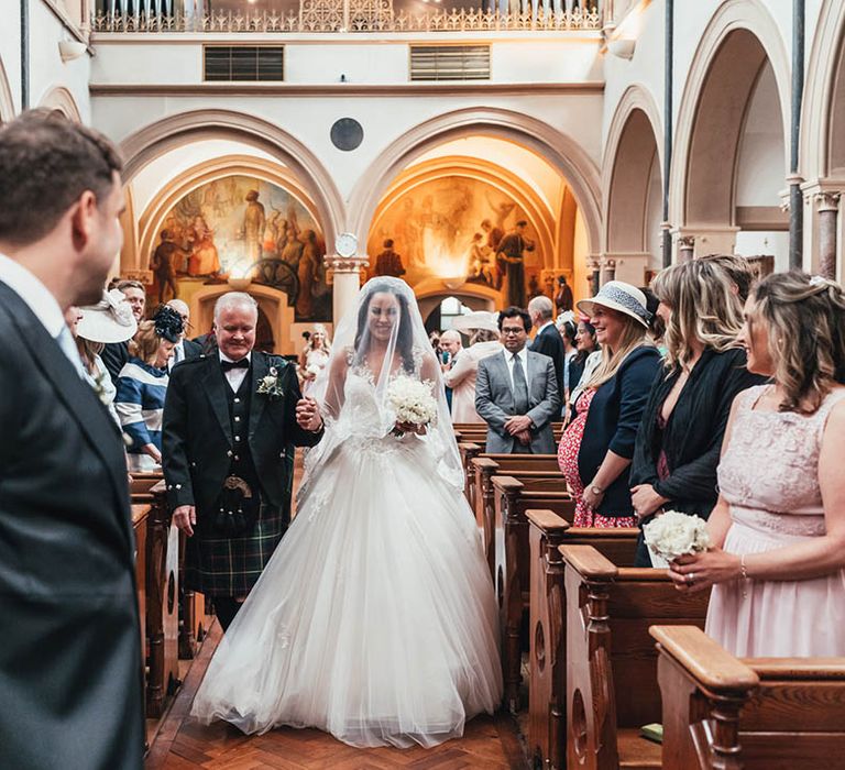 Father of the bride intertwines hands with bride and walks her down the aisle with veil to the groom at the altar 