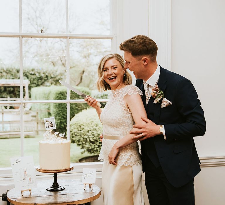 Bride & groom laugh as they cut their white frosted wedding cake complete with floral cake topper and picture 