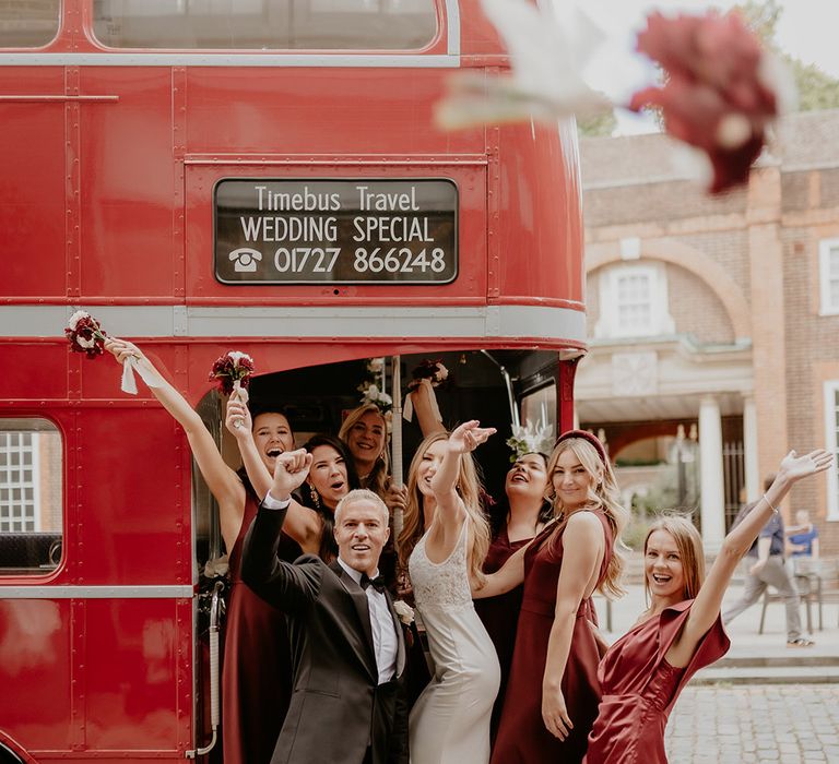 Groom in blacktop with the bride and the bridal party in dark red bridesmaid dresses on their red double decker bus wedding transport 