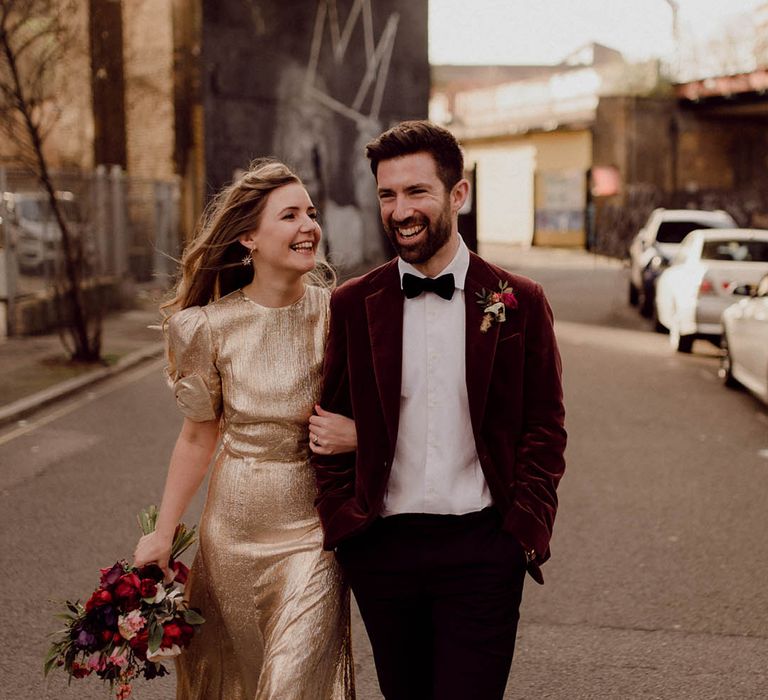 Bride wears gold wedding gown with bows to short sleeves whilst walking alongside her groom wearing a burgundy velvet blazer 