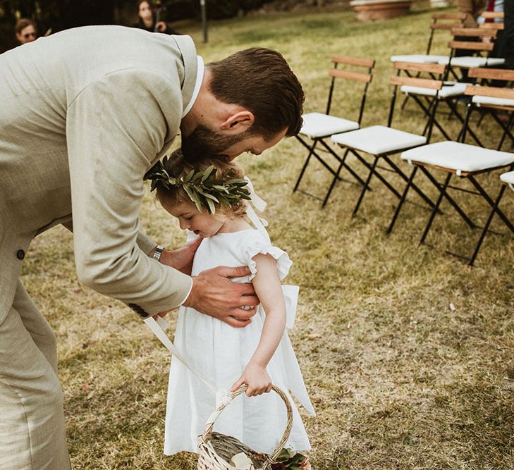 Groom kisses the head of flower girl who wears flower crown, white dress and carries wicker basket 