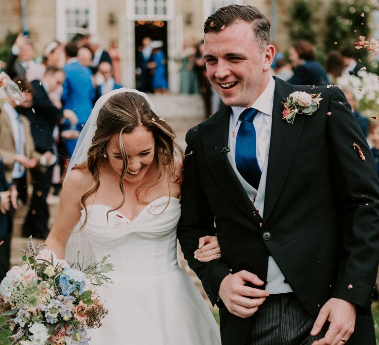 Bride wearing sweetheart neckline wedding dress & groom wearing morning suit walk through colourful confetti at Cowdray House