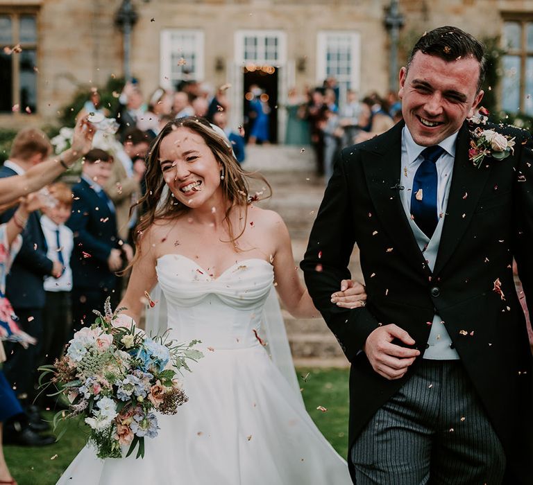 Bride wears sweetheart neckline wedding dress with ruched front whilst walking through confetti with her groom