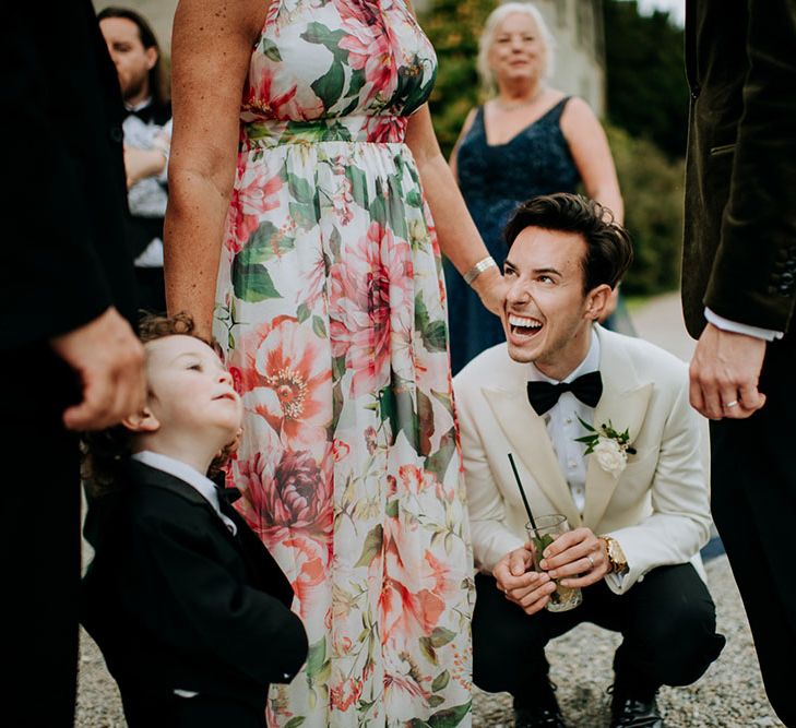 Groom wearing white dinner jacket and white floral rose buttonhole kneels down to little boy outdoors at Birdsall House