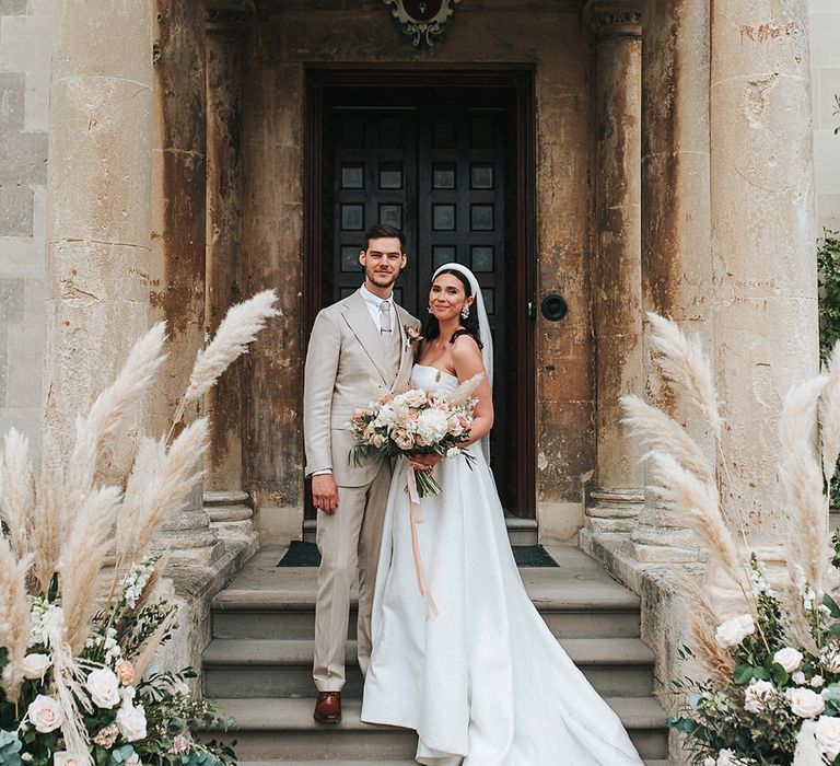 Bride in a strapless wedding gown holding blush pink bouquet and a chunky white headband with the groom in a neutral suit with pampas grass floral arrangements