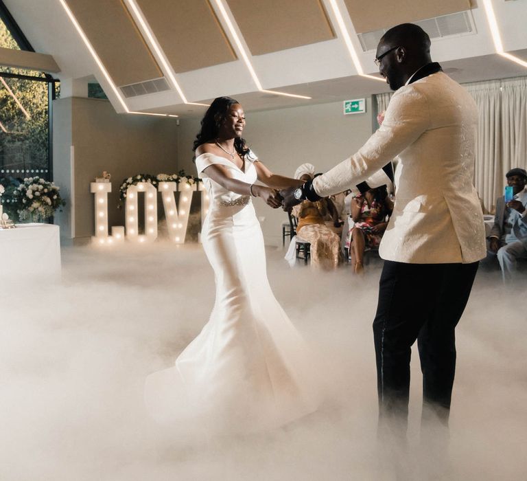 Bride & groom dance with one another in front of white rose flower wall and neon sign as mist covers the dance floor and wedding guests watch on