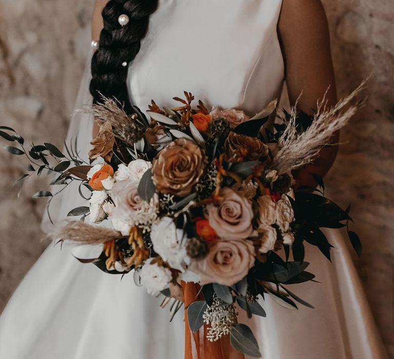 Black bride wears her hair in intricate braided styling complete with pearl hair accessories whilst wearing classic satin wedding dress and holding dried floral bouquet with Autumnal colour scheme