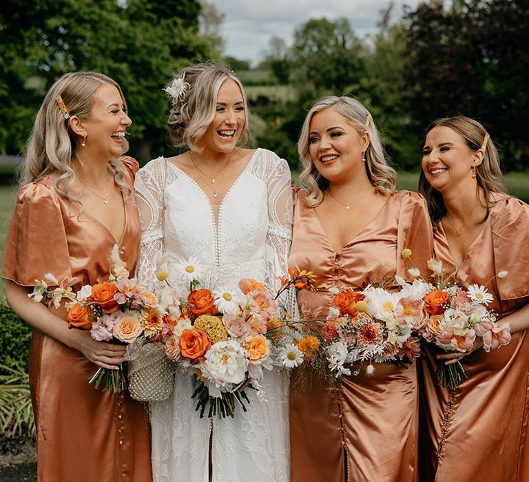 Bridal party with bridesmaids in peach satin dresses with bride in boho lace wedding dress holding orange bouquets