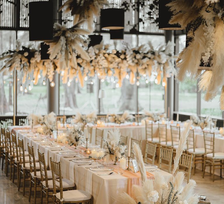 Pampas grass hangs above banquet tables complete with pastel pink fabric table runner complete with small dried floral centrepieces 