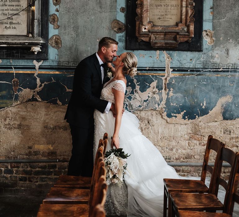 Bride & groom embrace in front of rustic wall at The Asylum on their wedding day 