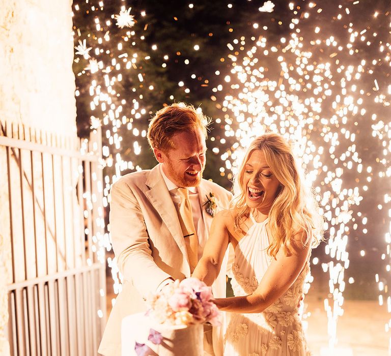 Bride and groom cut their three-tier rustic wedding cake decorated with purple flowers as fireworks explode