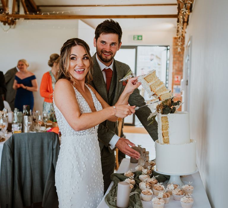 Bride in sleeveless Made With Love Bridal dress and groom in grey suit cutting the three tiered white buttercream wedding cake with burnt orange roses decorations