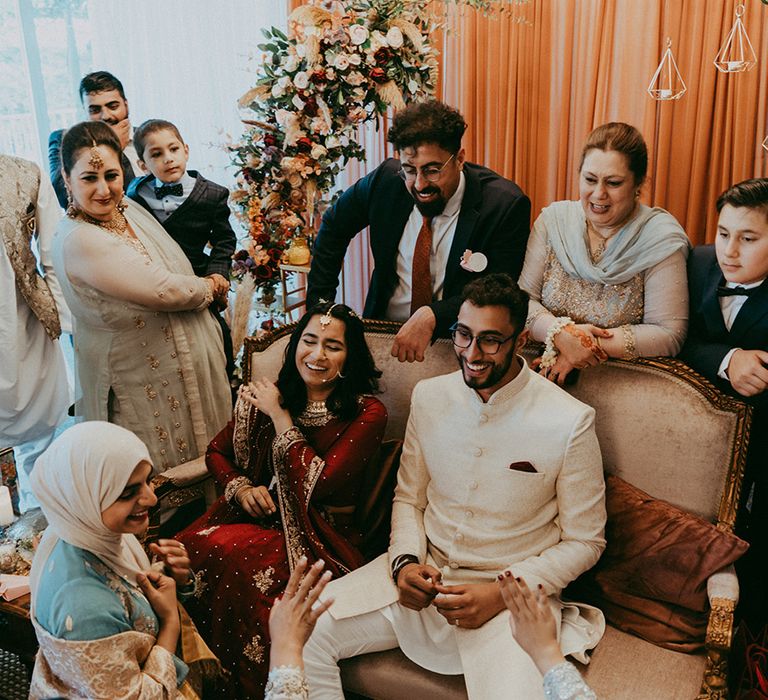 Bride & groom sit in front of orange curtain surrounded by wedding guests during Nikkah ceremony 