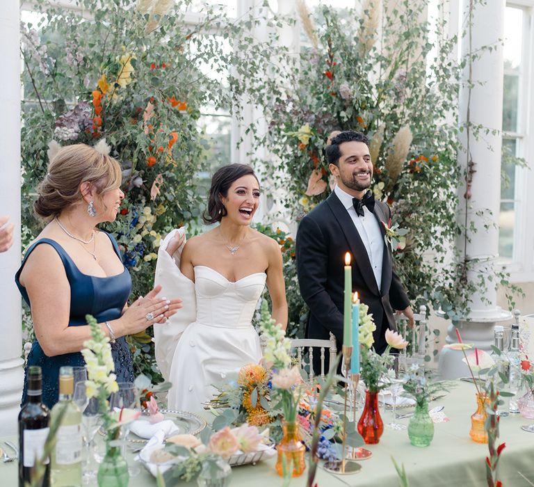 Bride & groom stand in front of brightly coloured floral installation complete with small colourful vases on pastel green tablecloth 