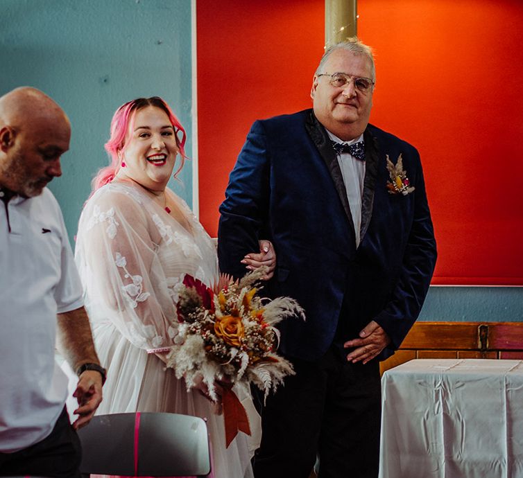 Bride in corset top wedding dress with leaf detailed lace overlay, pink stitching around the waist and red gemstone necklace holding autumnal coloured bouquet with white pampas grass and an orange ribbon and Father of the bride in midnight blue blazer with galaxy patterned bowtie and autumnal boutonniere