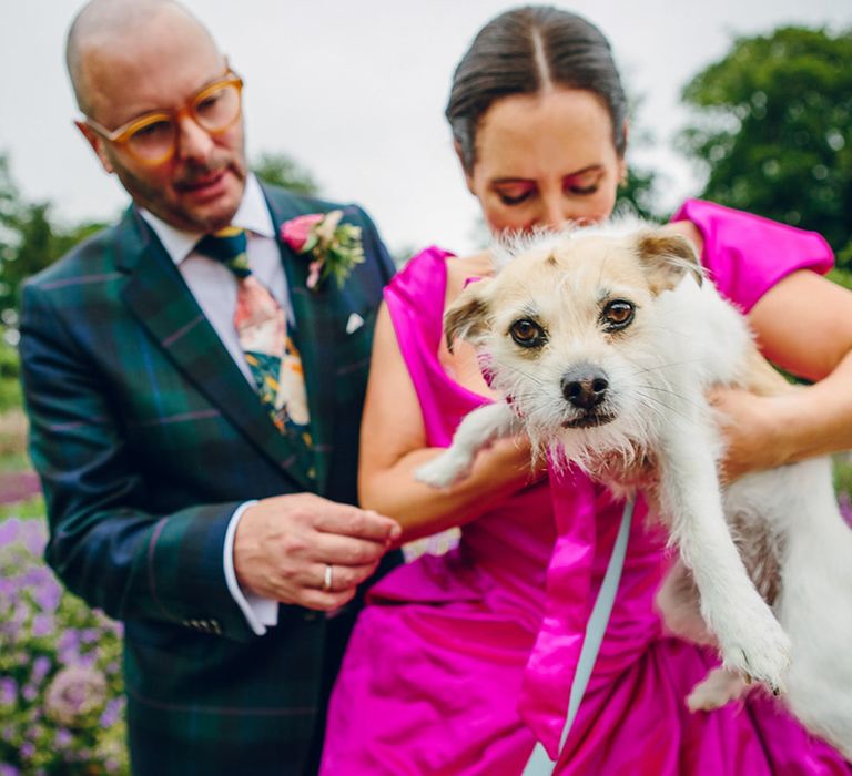 Groom in dark forest green tartan suit with patterned tie, white pocket square and pink and green boutonniere and bride in off the shoulder front ruching pink wedding dress holding Dolly the dog with a large satin pink bow 