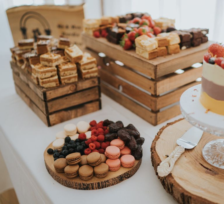 Wooden crate with a variety of cakes and fruit, wooden slate with dates, raspberries and macaroons, and wooden slate with single tier buttercream vegan wedding cake 