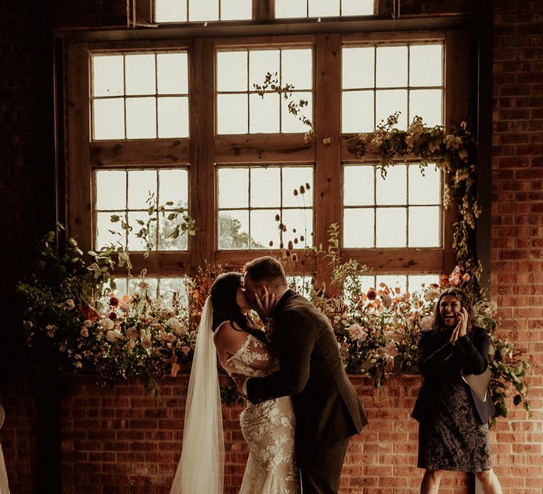 Bride in off the shoulder Abigail Westrup bridal gown with long, sheer sleeve and lace detailing and cathedral length veil kissing groom in dark suit at The Giraffe Shed 