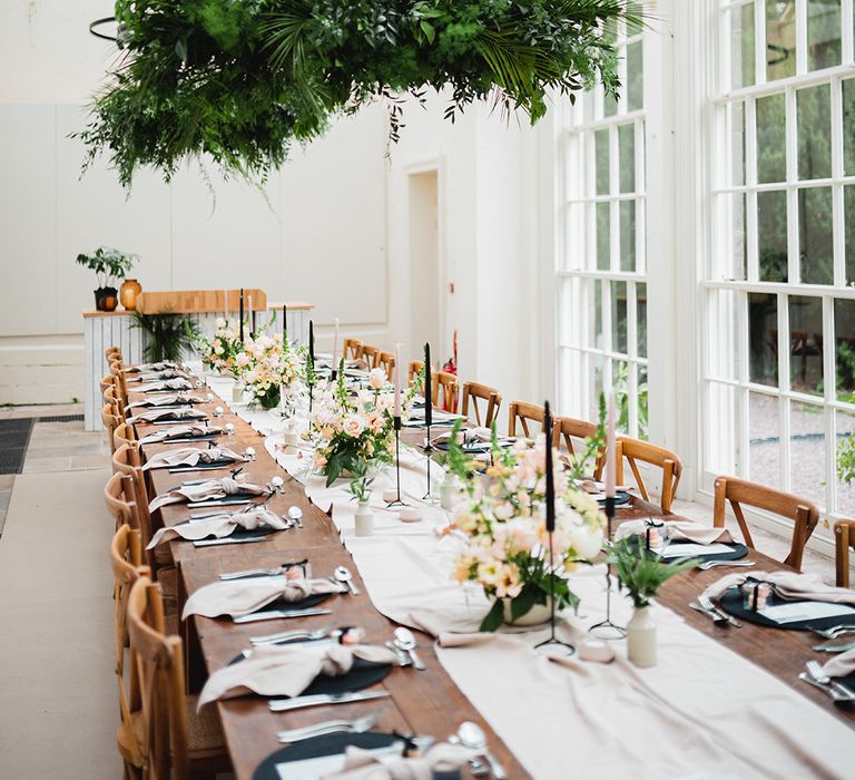 Reception room of The Orangery Ingestre wedding venue with neutral off-white table runners, rose and foliage floral arrangements, pink and black tapered candles, black candlestick holders and suspended foliage decorations