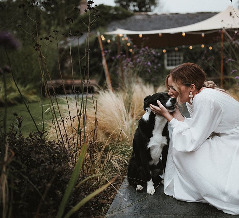 Bride in silk floaty wedding dress kisses dog on the day of her wedding outdoors 