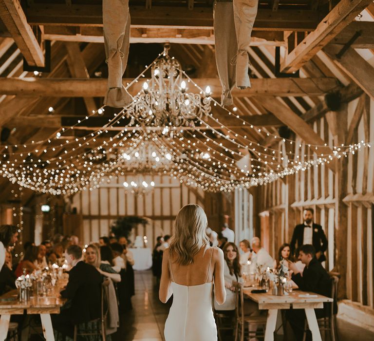 Fairy lights decorate Southend Barns wedding venue as the bride turns to face the guests isttingon long banquet tables at the cosy rustic wedding 