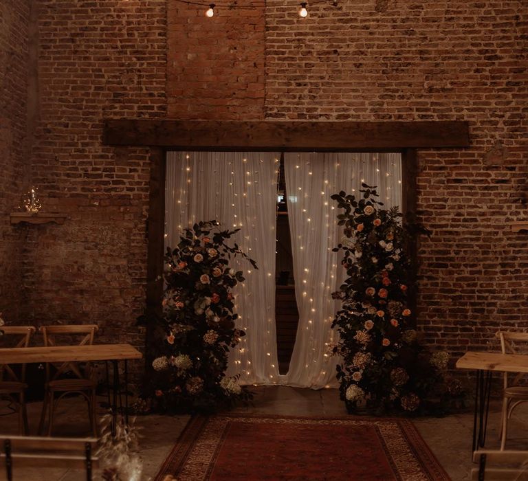 Rustic barn wedding with pale pink roses on a flower column in front of white wedding drapes and fairy lights decorating the altar 