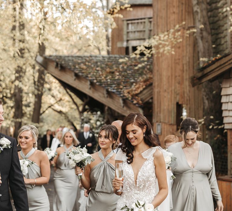 Bridesmaids in satin sage green bridesmaid dresses with the bride in a fitted lace wedding dress with bow straps and white orchid and rose cascading bouquet