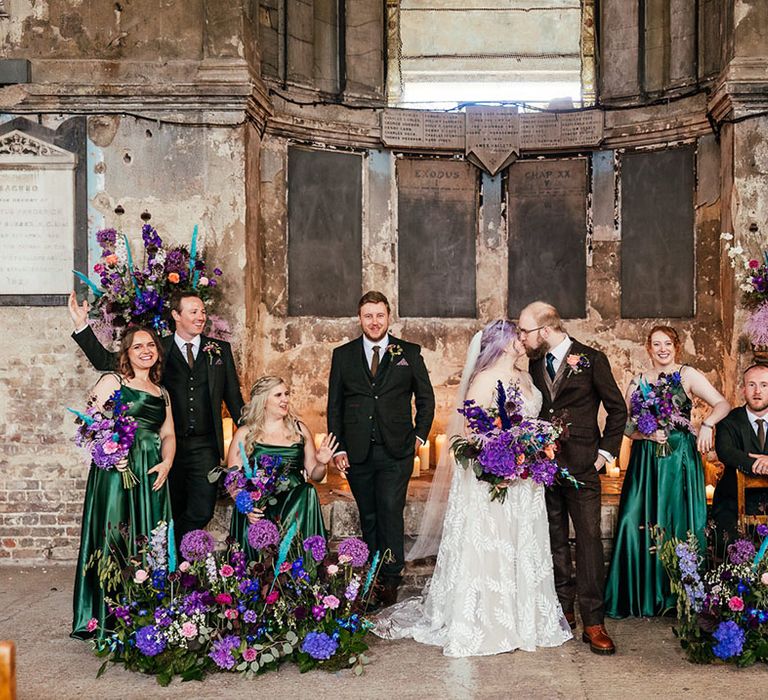 Bride in dainty leaf appliqué sheer Evie Young bridal gown and cathedral length veil kissing groom in mocha brown textured suit with deep green tie and pink and purple boutonniere, holding bright purple bridal bouquet with purple hydrangeas, pink garden roses, purple orchids, lilac carnations, dried flowers and peacock blue feathers standing with bridesmaids in tie strap forest green bridesmaid dresses and groomsmen in dark three piece textured suits with dried flower boutonnieres, surrounded by large purple, blue and pink floral displays 